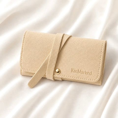 A beige faux-leather jewelry organizer with a strap closure and a gold snap button, featuring the brand name 'KinMarked.' embossed on the front, resting on a white satin fabric background. KinMarked Personalized Jewelry Travel Roll for Jewelry Gifts