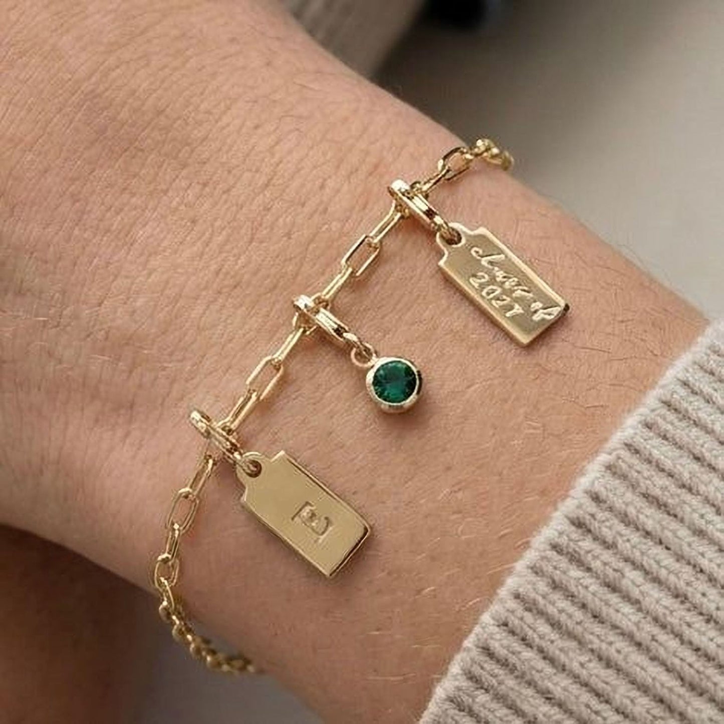 A gold bracelet with a charm and a tag attached to it, worn on a person's wrist.