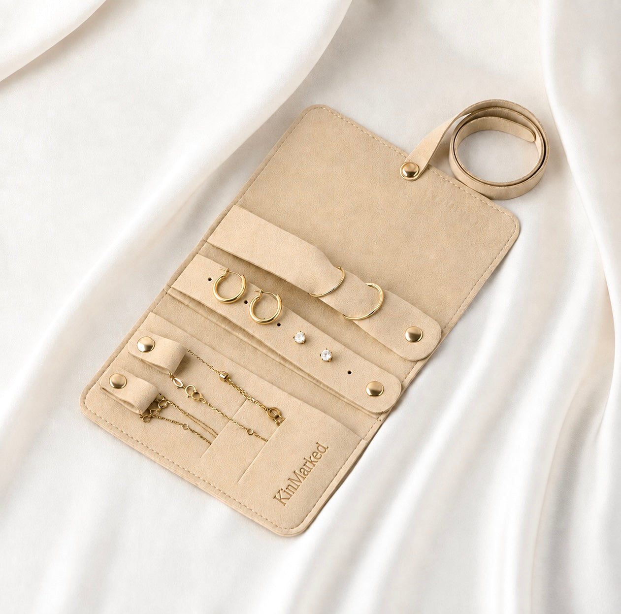 A beige faux-leather jewelry organizer roll-up pouch with gold-tone hardware, shown open on draped white fabric. The interior includes slots for rings, earrings, pins, and chains, with the brand name 'KinMarked' printed on the bottom. KinMarked Weekender Travel Roll for Personalized Jewelry Gifts