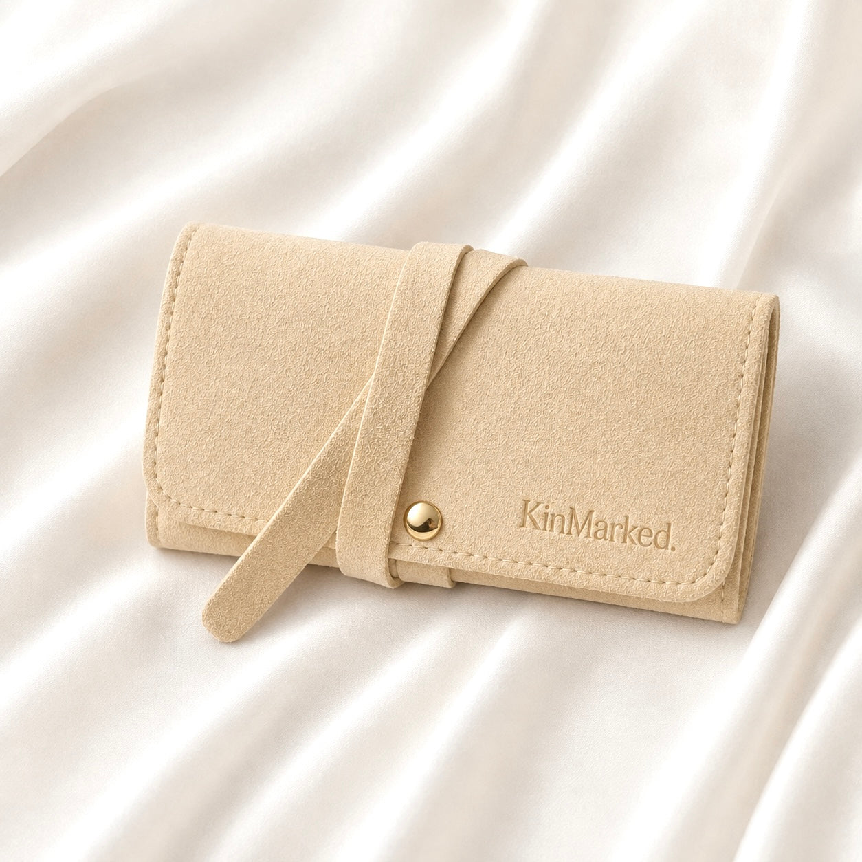 A beige faux-leather jewelry organizer with a strap closure and a gold snap button, featuring the brand name 'KinMarked.' embossed on the front, resting on a white satin fabric background. KinMarked Personalized Jewelry Travel Roll for Jewelry Gifts