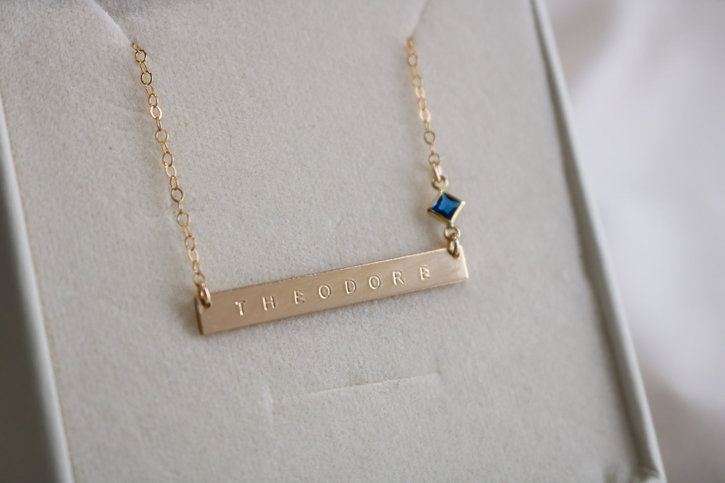 Gold name necklace with engraved 'THEODORE' and blue gemstone accent.