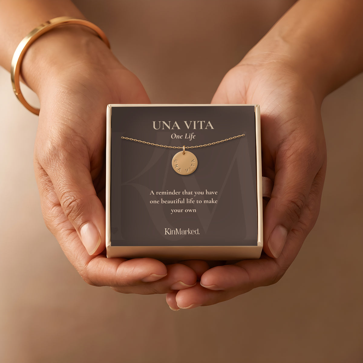 Hands holding a KinMarked gift box with the Una Vita hand-stamped 13 mm 14k gold-filled disc necklace on its One Life meaning card