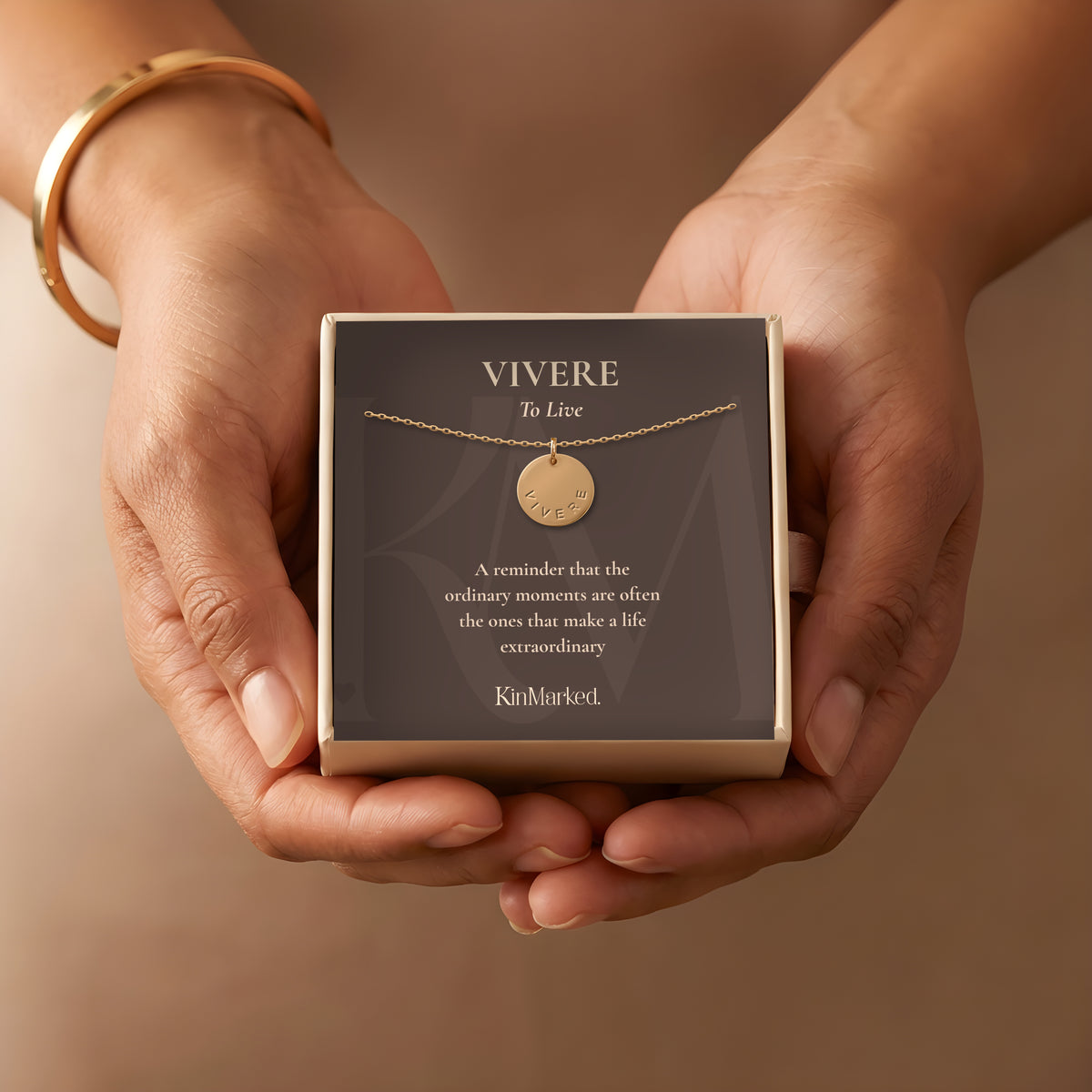 Hands holding a KinMarked gift box with the Vivere hand-stamped 13 mm 14k gold-filled disc necklace on its To Live meaning card